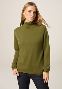 Maglione a collo alto verde oliva, texture morbida, maniche lunghe, vestibilità rilassata, indossato con jeans scuri. Nota per il design semplice e pulito.