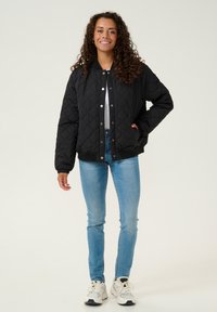 Veste bomber noire matelassée avec un col arrondi, fermeture à boutons-pression et poignets côtelés, portée avec un jean skinny bleu clair et des baskets blanches.