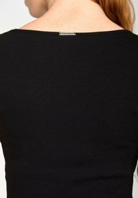 Close-up van de bovenrug van een persoon die een zwarte geribbelde top draagt met een klein metalen label 'Vive Maria' bij de halslijn.