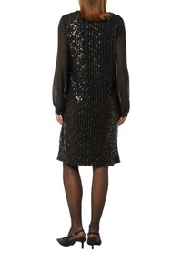 Kobieta stoi tyłem do kamery, ubrana w czarną cekinową sukienkę bez rękawów na przezroczystej bluzce z długim rękawem oraz czarnych szpilkach.