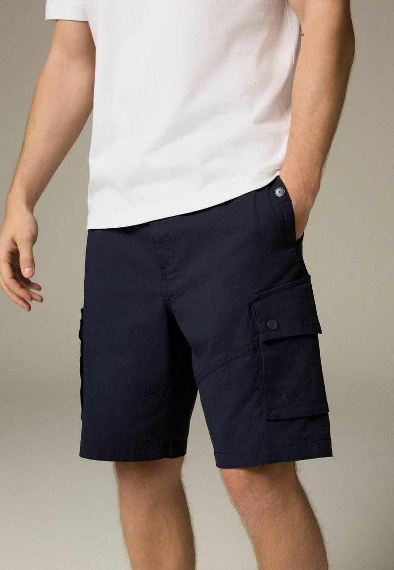 Pantaloni scurți de tip cargo navy, confecționați dintr-un material texturat, având buzunare laterale cu clapete, accente cusute și închidere cu buton la talie.