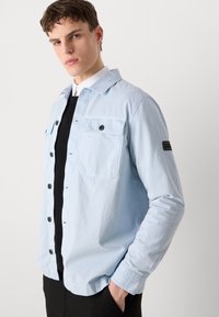 Hele sinine särk-jakk nupuga esiküljel, rinnatasku ja mustad nupud. Omab musta käisepleki ja lõdva istuvust.