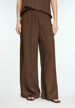 Kobieta w luźnych, brązowych spodniach o szerokich nogawkach, połączonych z dopasowaną brązową bluzką i czarnymi sandałami na kwadratowym obcasie z odkrytymi palcami.