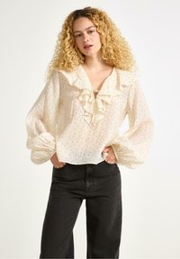 Hvid bluse med flæsekrave og sorte prikker, med lange puff ærmer og bundet halsudskæring, parret med sorte højtaljede jeans.