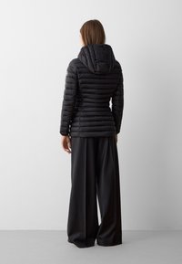 Giacca a vento nera con design trapuntato e cappuccio, vista da dietro, abbinata a pantaloni neri a gamba larga. Texture liscia, dettagli minimi.