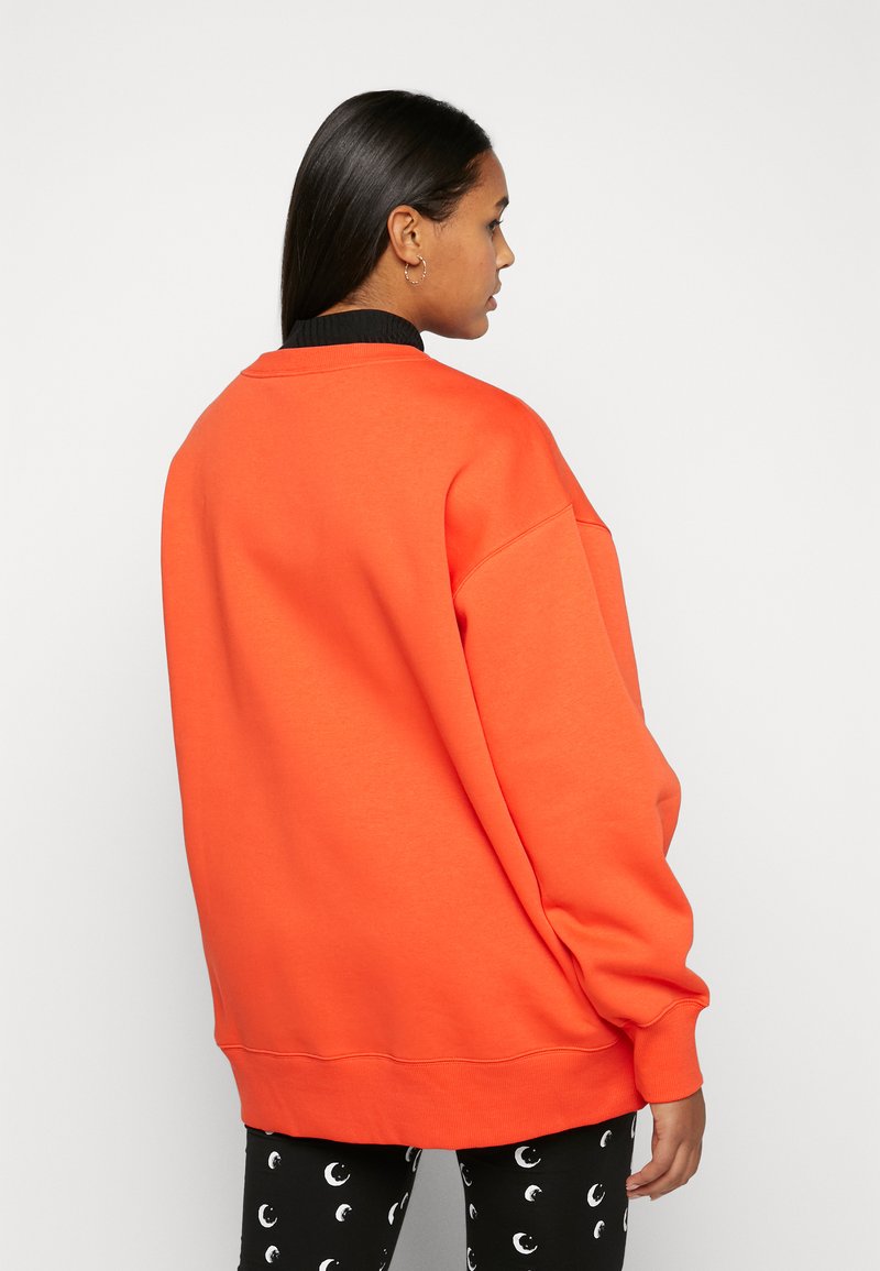 Sweatshirt orange surdimensionné avec un col rond et un col côtelé noir, associé à un pantalon noir avec des motifs de croissant de lune blancs.