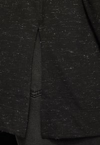 Schwarzstoffkleidung mit einer strukturierten Oberfläche, die weiße Sprenkel und einen seitlichen Schlitz aufweist. Nahaufnahme, die die Stichdetails hervorhebt.