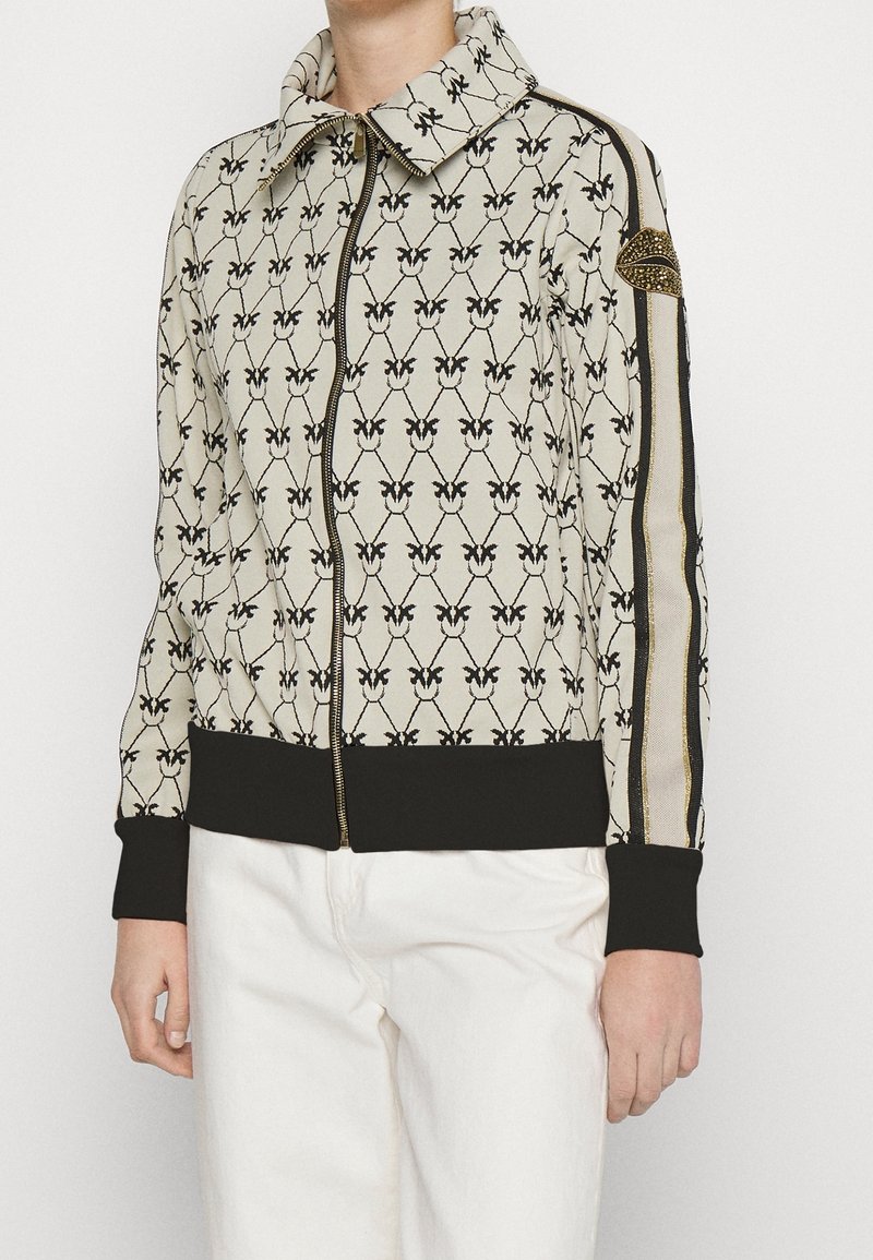 Chaqueta bomber texturizada con un fondo beige y un patrón de mariposas negras, ribetes y puños negros, detalles dorados en las mangas y cierre de cremallera.