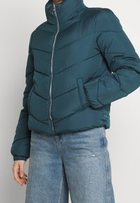 Turkis puff-jakke med høj krave, lynlås foran og diagonalt quiltet mønster. Har elastiske manchetter og sidelommer. Bruges med lyseblå jeans.