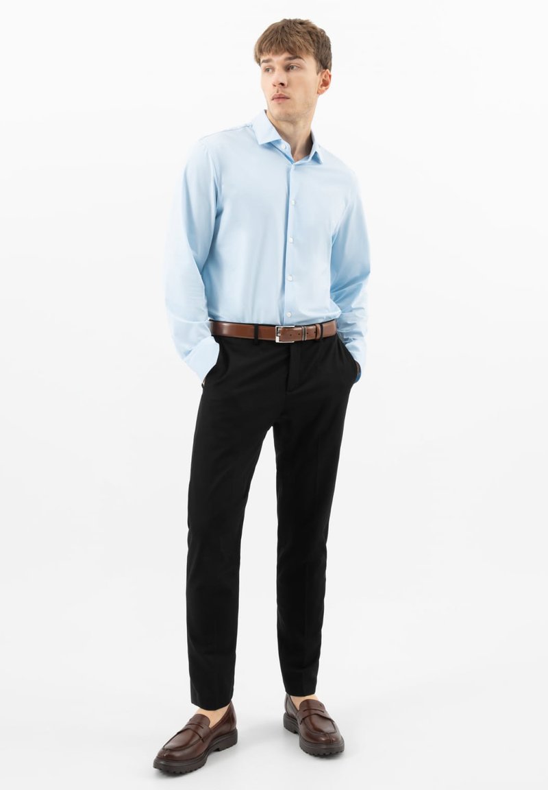 Giovane uomo in piedi con le mani nelle tasche, indossa una camicia azzurra, pantaloni neri, cintura marrone e mocassini marroni su uno sfondo bianco.