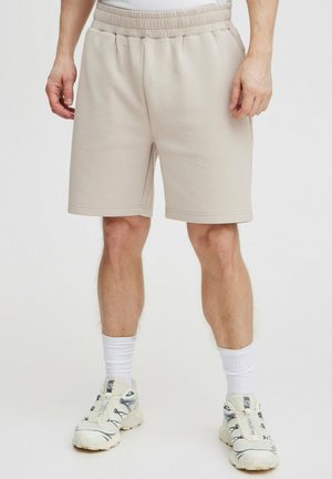 Shorts en tricot beige avec une taille élastique, des poches latérales et une coupe décontractée. Portés avec des chaussettes blanches et des chaussures de sport de couleur claire.