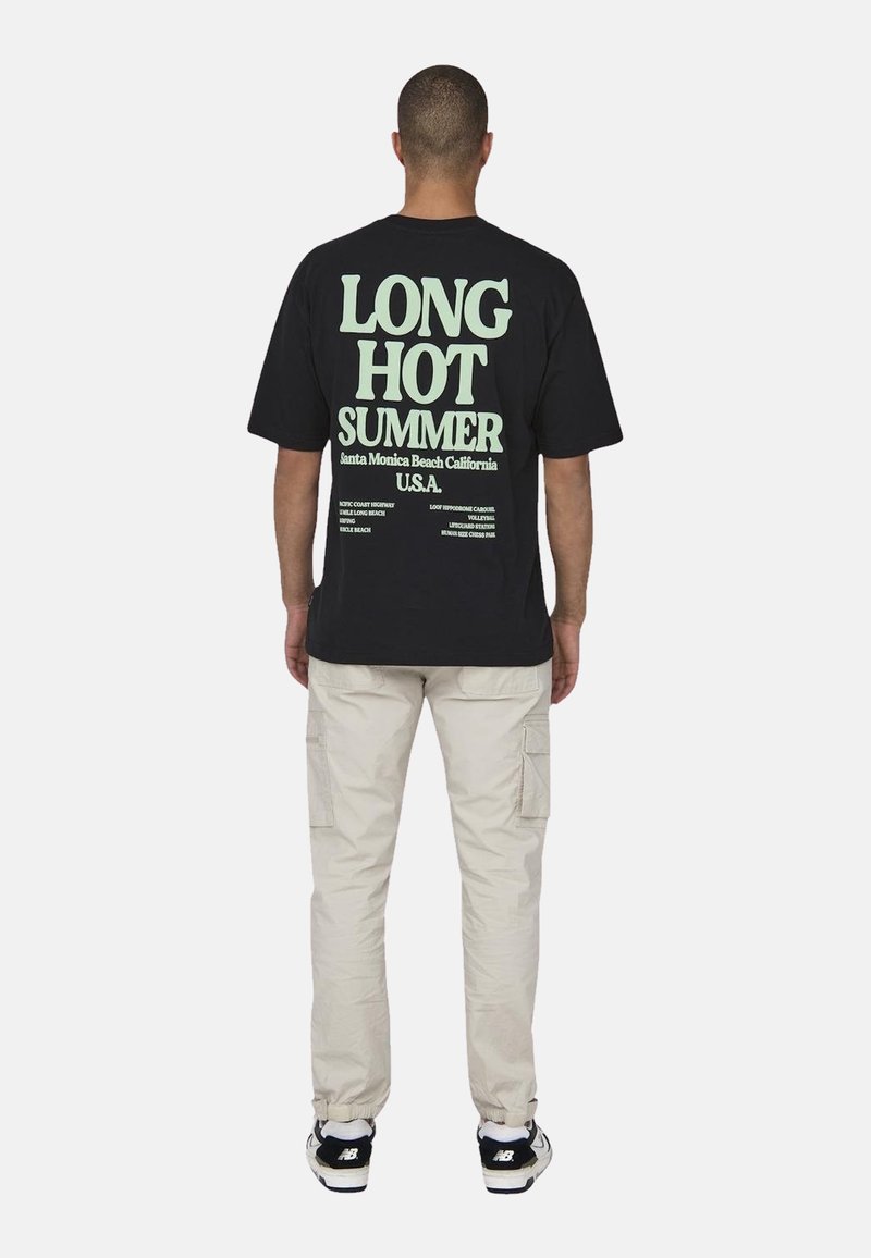 Czarny t-shirt z dużym zielonym napisem "DŁUGIE GORĄCE LATO" na plecach. W komplecie z jasnobeżowymi spodniami cargo i czarnymi trampkami.