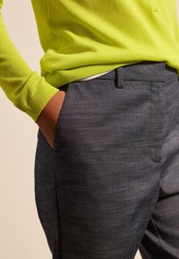 Neongrüner Strickpullover kombiniert mit dunkelgrauen, taillierten Hosen; die Hosen weisen ein dezentes strukturiertes Muster und Seitentaschen auf.