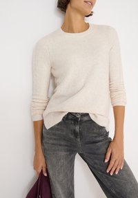 Hellbeige Pullover mit Rundhalsausschnitt und geripptem Saum, kombiniert mit dunkelgrauen Jeans. Der Pullover hat subtile strukturierte Details an den Ärmeln.