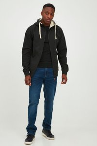 Black zip-up hoodie with cream drawstrings, worn over a black shirt, paired with dark blue jeans and black sneakers. Smooth fabric, casual fit.