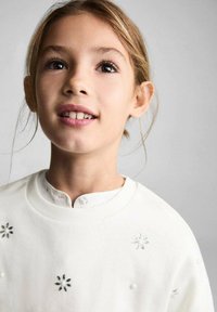 Vit sweatshirts med blomsterdekorationer och pärlprydnader, med en ribbad halsringning och en lager av vit skjortkrage under.
