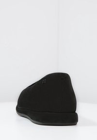 Scarpa nera slip-on con una texture liscia e opaca, forma arrotondata e un discreto marchio sul tallone. Progettata per il comfort con una suola piatta.