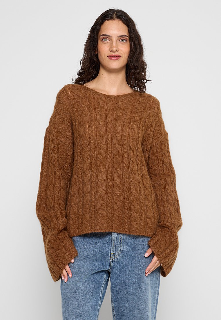 Brązowy sweter dziany z wzorem w warkocze, szerokimi rękawami i dekoltem w łódkę, w połączeniu z niebieskimi dżinsami. Miękka faktura i luźny krój.