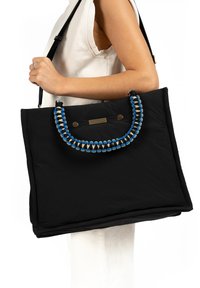 Zwarte tote bag met een gewatteerde textuur, voorzien van een gevlochten handvat met blauwe en gouden accenten. Drukknoopsluiting en decoratieve hardware detail.