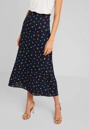 Femme portant une jupe midi bleu marine avec de petites impressions florales colorées, associée à un haut blanc et des sandales à talons hauts dorées.