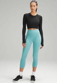 Schwarzes Crop-Top mit langen Ärmeln, kombiniert mit türkisfarbenen, hochgeschnittenen Leggings. Das Modell trägt schwarze Sneakers mit weißen Akzenten. Glatte Texturen.