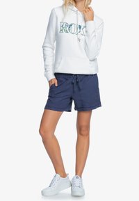 Bílá mikina s kapucí a nápisem "ROXY" v zeleném květinovém vzoru, námořnické šortky s elastickým pasem a bílé tenisky s texturovanou podešví.