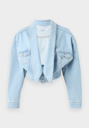 Kobieta ubrana w jasnoniebieską, oversize kurtkę jeansową oraz jeansy z wysokim stanem i szerokimi nogawkami, biała bluzka i czarne, szpiczaste buty, stojąca na białym tle.