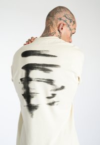 Maglietta color crema con un ampio design astratto di pennellate nere sul retro. Realizzata in morbido cotone con una vestibilità comoda.
