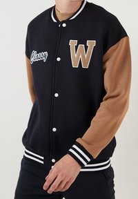 Mörkblå och brun varsityjacka med vita detaljer, med en stor "W"-emblem, knappar och ribbade muddar. Mjuk tyg med kontrasterande ärmar.