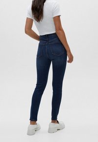 Kvinna står med ryggen mot kameran iförd mörkblå skinny jeans, vit kortärmad t-shirt och vita kraftiga plattformsskor.