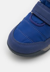 Modrá sportovní obuv s polstrovaným svrškem, suchým zipem, texturovaným materiálem a gumovou podrážkou, se švy detailů a kulatou špičkou.