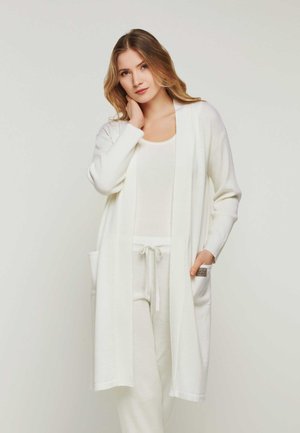 Langer, weißer Cardigan aus weichem Material, mit offenem Vorderteil, weit geschnittenen Ärmeln und zwei vorderen Taschen. Kombiniert mit einem weißen Oberteil und Hosen.