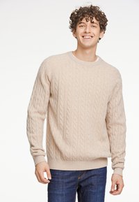 Beiger Kabelstrickpullover mit Rundhalsausschnitt, gerippten Bündchen und Saum, weicher Textur, über einem weißen Hemd getragen, kombiniert mit blauen Jeans.