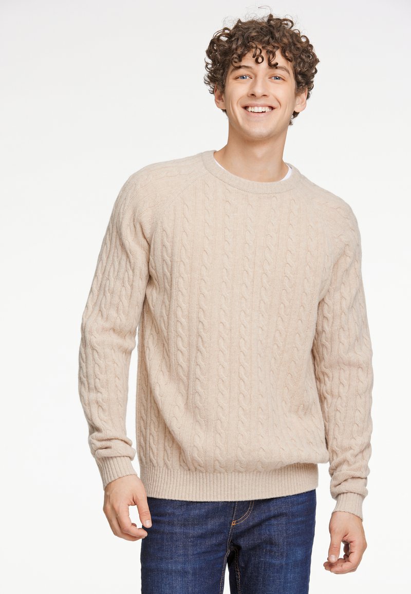 Beiger Kabelstrickpullover mit Rundhalsausschnitt, gerippten Bündchen und Saum, weicher Textur, über einem weißen Hemd getragen, kombiniert mit blauen Jeans.