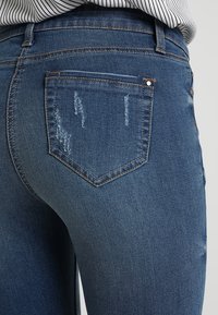 Donkerblauwe denim jeans met een versleten textuur, achterzakken met stikseldetails en een middelhoge taille voor een aangesloten silhouet.
