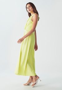 Kelio ilgio fluorescencinės geltonos spalvos suknelė su sukimo priekyje detale, turinti plačias petnešas ir laisvą siluetą. Derinama su baltomis šlepetėmis.