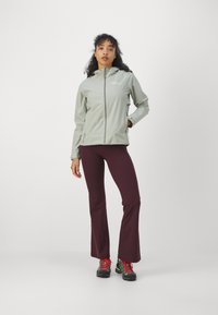 Lichtgroene zip-up jas met capuchon, gecombineerd met kastanjebruine uitlopende broek. Kenmerken zijn een zachte textuur en sportieve zwarte wandelschoenen.