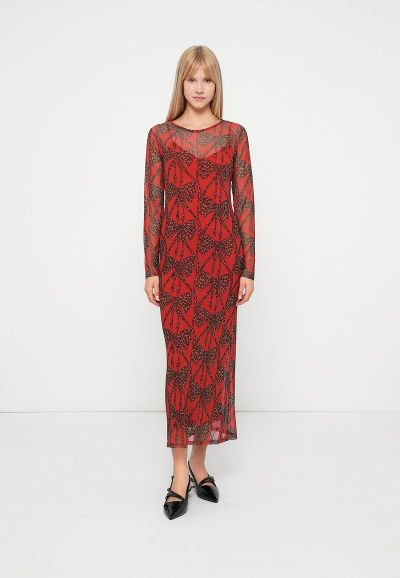 Długa czerwona sukienka z czarnym wzorem liści, przezroczysty materiał, okrągły dekolt i długie rękawy, połączona z czarnymi butami na spiczastym obcasie.
