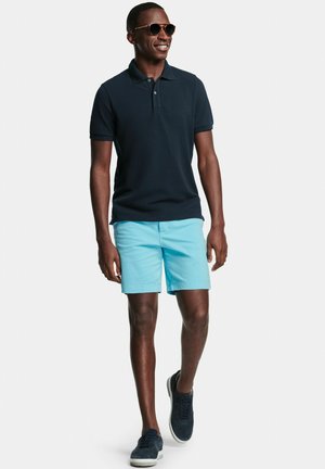 Man met een zonnebril, een donkerblauw poloshirt, lichtblauwe shorts en donkere sneakers die tegen een effen witte achtergrond loopt.