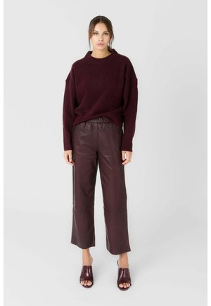 Femme portant un pull bordeaux, un pantalon en cuir large assorti et des mules à talons ouvertes, debout devant un fond clair uni.