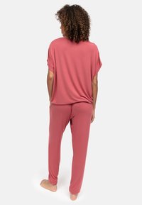 Person mit lockigem Haar steht barfuß mit dem Rücken zur Kamera, trägt ein lockeres rosa kurzärmeliges Oberteil und passende Hosen vor weißem Hintergrund.