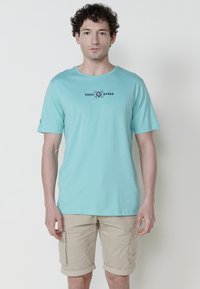 Türkises Baumwoll-T-Shirt mit Rundhalsausschnitt und kurzen Ärmeln, mit einem dunkelblauen "Kashi Brand"-Logo auf der Brust. Getragen mit beigen Cargo-Shorts.