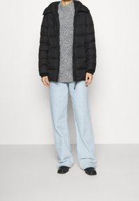 Sort pufferjakke med hætte, quiltet design, båret over en grå tekstureret sweater, parret med lyseblå vidde bukser og sorte sko.