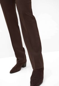 Personne portant un pantalon ajusté marron foncé et des bottines en daim marron foncé assorties à talons carrés sur fond blanc.