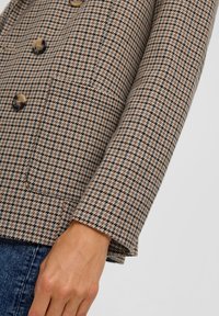 Karierter Blazer in Beige und Schwarz, mit einem doppelt geknöpften Design, großen Knöpfen und einer Fronttasche. Strukturierter Stoff und taillierte Passform.