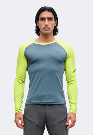 Uomo con capelli neri che indossa una maglietta sportiva aderente a maniche lunghe con corpo blu e maniche giallo brillante, in piedi davanti a uno sfondo semplice.
