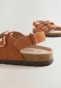 Braune Wildleder-Sandalen mit rosa geflochtenen Akzenten, verstellbaren Riemen und floralen Details auf der Oberseite. Korkfußbett und strukturierte Sohle.
