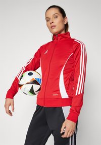 Red athletic jacket with white stripes and zip closure, paired with black pants. A patterned soccer ball is held under the arm.