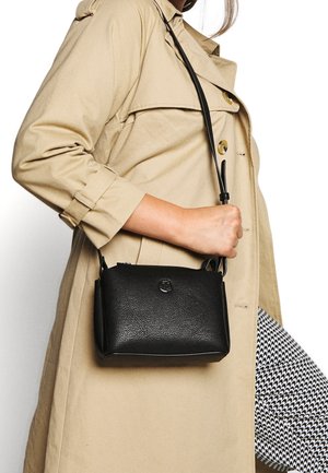 Femme en trench-coat beige tenant un sac bandoulière en cuir noir, portant un pantalon pied-de-poule noir et blanc.