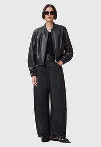 Giacca bomber nera in pelle con polsini a costine, abbinata a pantaloni neri larghi. Il modello indossa occhiali da sole e una collana a catena.
